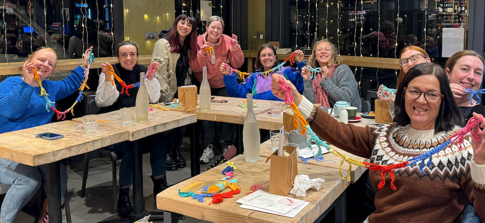 Group of people sitting around a table making crochet bow garlands in a cozy indoor cafe setting.